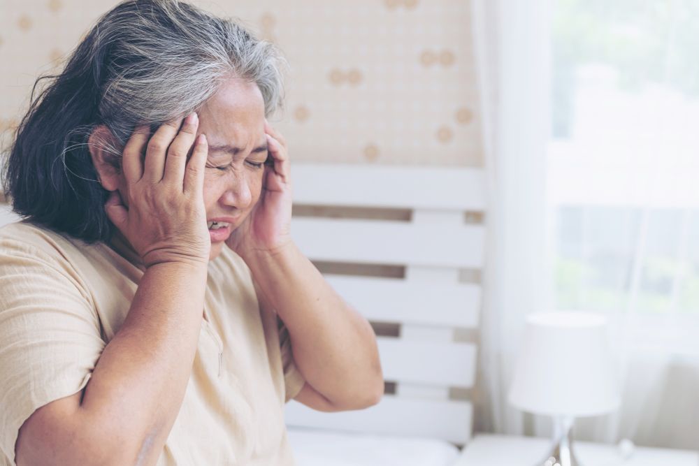 An older woman stands in a bedroom, holding her head with both hands and appearing distressed, perhaps in need of a chiropractor's touch to alleviate the relentless headache.