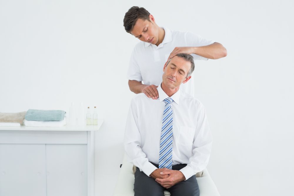 In a bright room, a man in a white shirt and tie sits while another man in white performs a neck massage, working his skills as a chiropractor to relieve the tension from an aching headache.