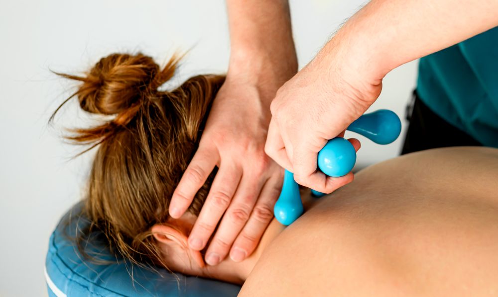 A person lies face down on a massage table, enjoying a soothing back massage with a blue tool, reminiscent of the relief often sought from a chiropractor for tension headaches.