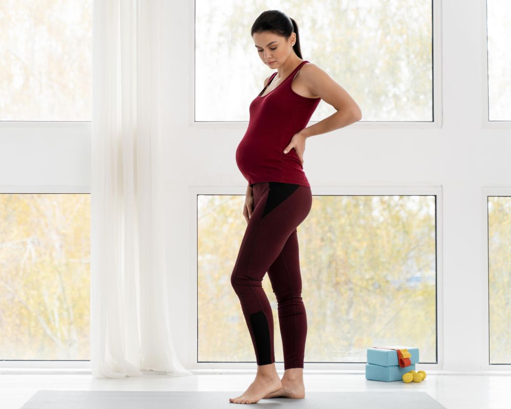 A pregnant woman in a red tank top and leggings stands barefoot in a bright room, looking down and holding her lower back, contemplating chiropractic care during pregnancy. A blue yoga mat and small weights are nearby.