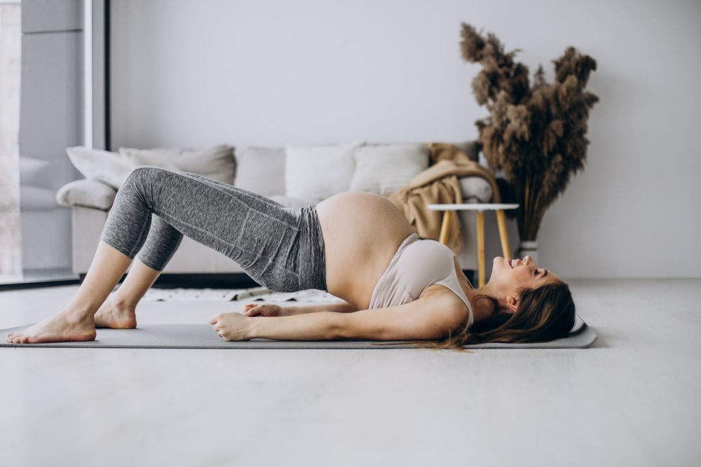 Pregnant woman performing a bridge exercise on a yoga mat in a living room, where the benefits of chiropractic during pregnancy can enhance comfort. A cozy sofa and decorative plants create a serene backdrop.